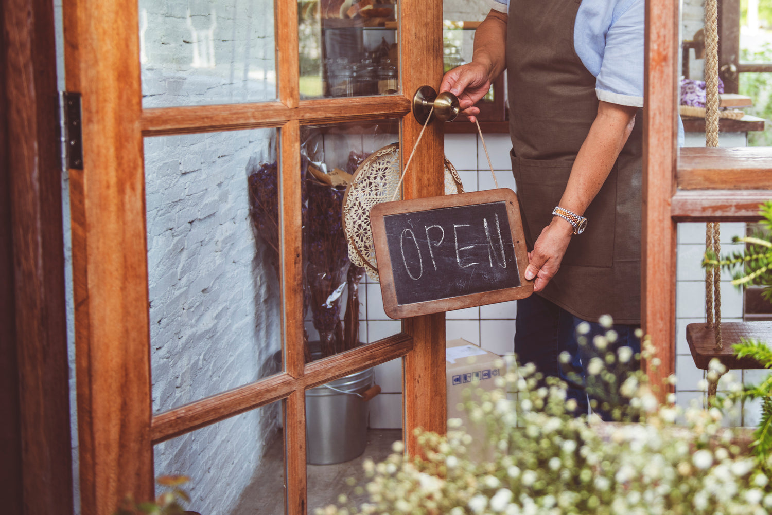 Person in an apron holding a 'Open' sign in front of a door with plants and decor inside.